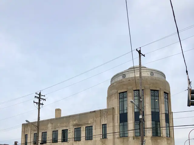 The image shows a white bus driving down a street lined with tall buildings, electric poles with...