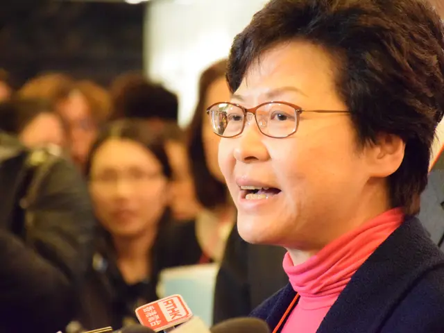 The image shows a woman wearing glasses and a pink scarf talking to reporters in front of a group...