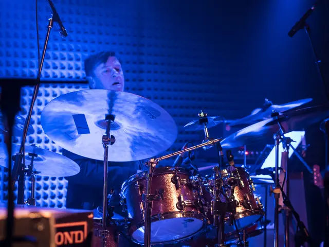 The image shows a man playing drums on stage at a concert, surrounded by mics with stands,...