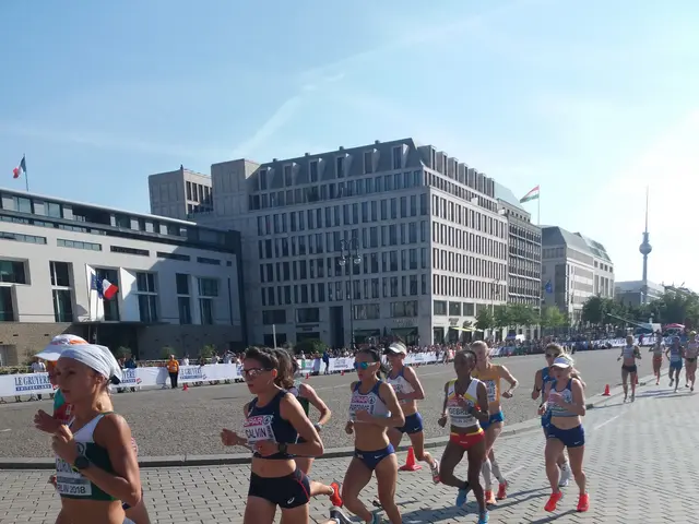 The image shows a group of women running in a marathon on a road surrounded by buildings, trees,...