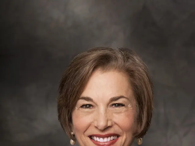 The image shows a woman in a blue suit smiling at the camera against a dark background. She appears...