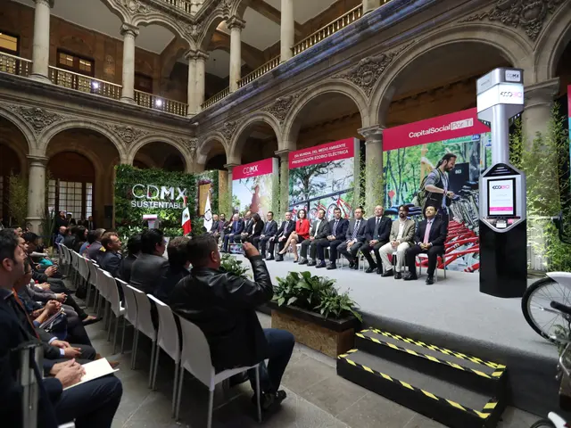 The image shows a group of people sitting in chairs in a building, with a man standing on a stage...