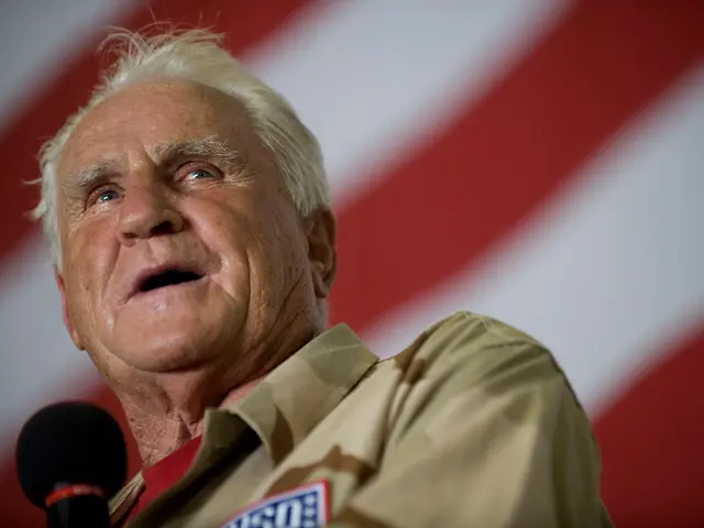 The image shows a man in a brown shirt speaking into a microphone in front of an American flag. He...