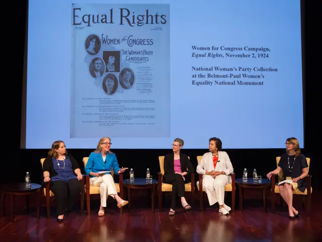 The image shows a group of women sitting on chairs on top of a stage, with tables in front of them...