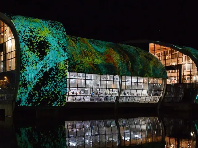 The image shows a building with a green roof lit up at night, surrounded by water and illuminated...