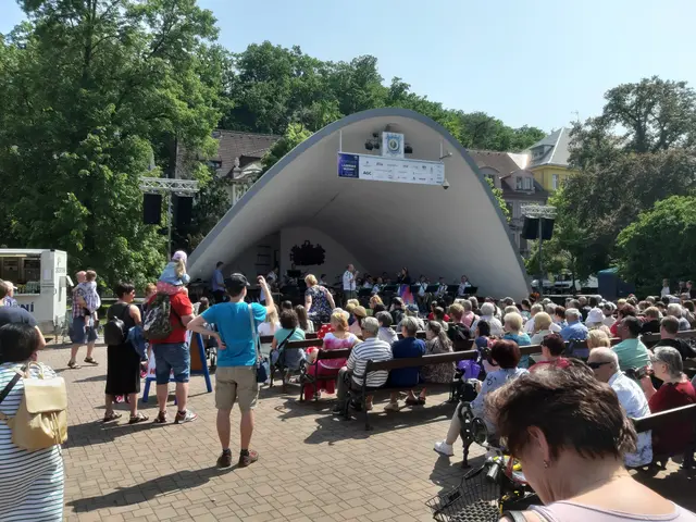 The image shows a crowd of people sitting on benches in front of a stage, with some people standing...