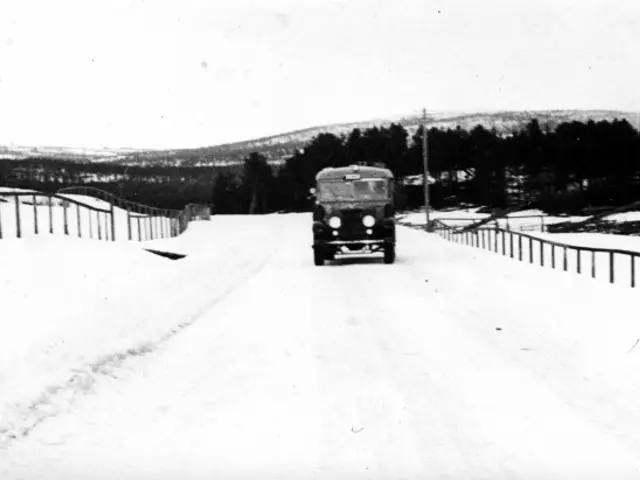 The image shows an old truck driving down a snowy road, surrounded by a fence on either side. In...
