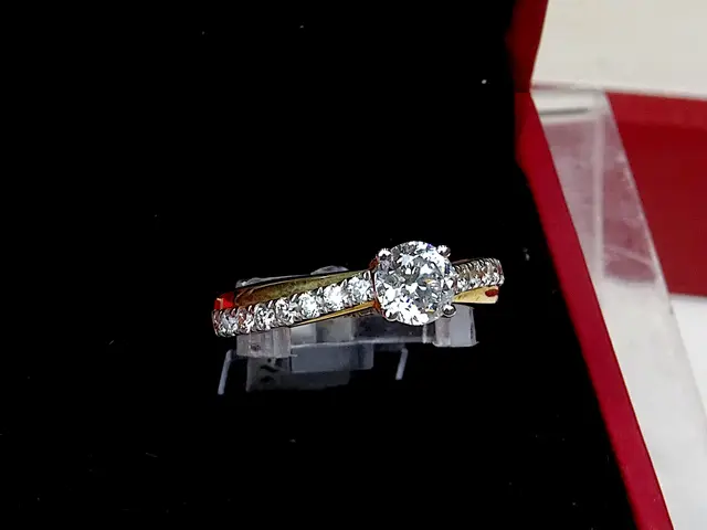 The image shows a diamond engagement ring in a red box on a table. The box is a deep red color and...