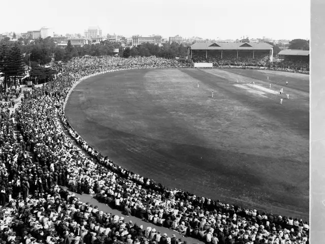 The image shows a black and white photo of a large crowd of people watching a cricket match in a...