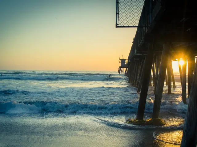 The image shows a pier on the beach at sunset with the sun setting in the background. The water is...