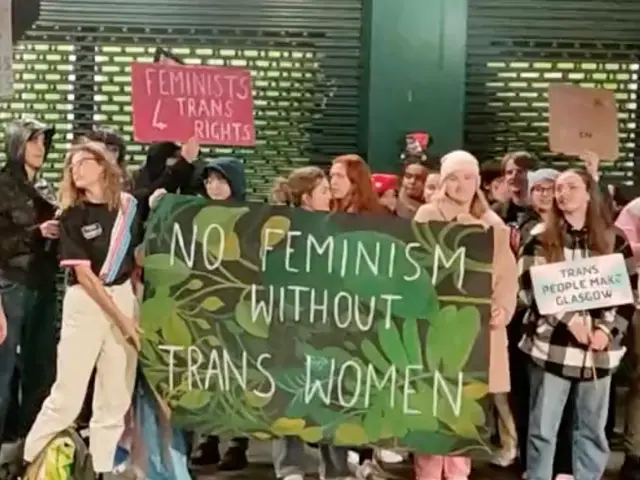 The image shows a group of people standing on the ground, holding a banner that reads "No Feminism...