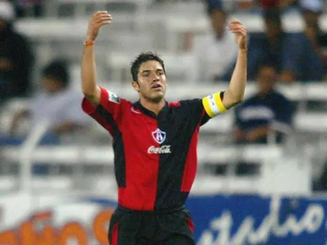 The image shows a soccer player in a red and black uniform running on a field with his arms...