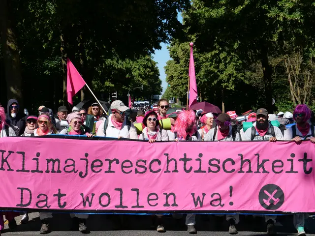 The image shows a group of people walking down a street, holding a pink banner with the words...