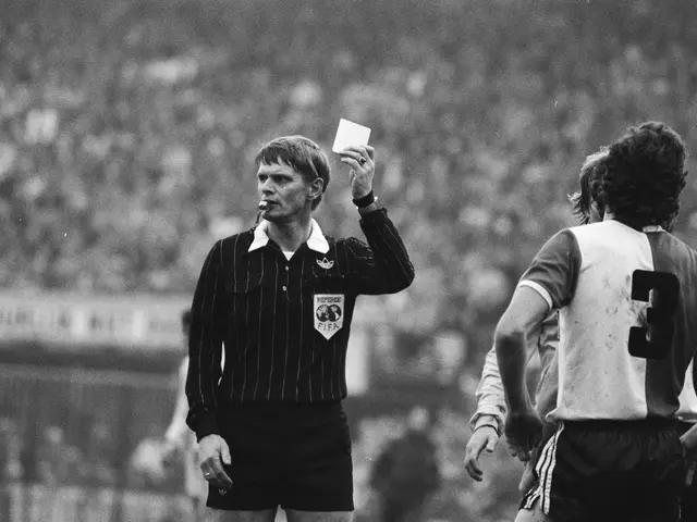 The image shows a referee holding a red card in front of a crowd of people, with a fence in the...