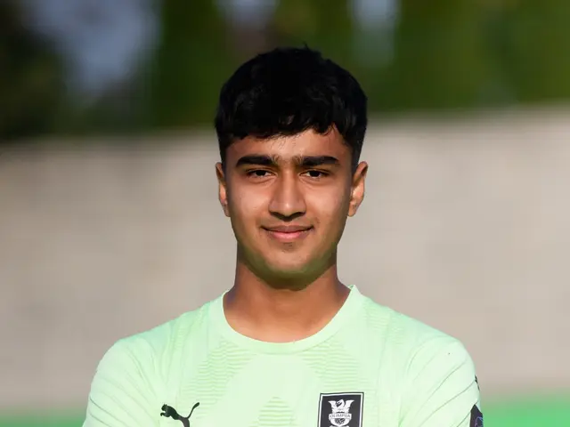 The image shows a young man in a green shirt standing on a soccer field with his arms crossed,...