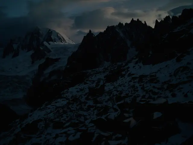 The image shows a snow-covered mountain range with a cloudy sky in the background. The mountains...