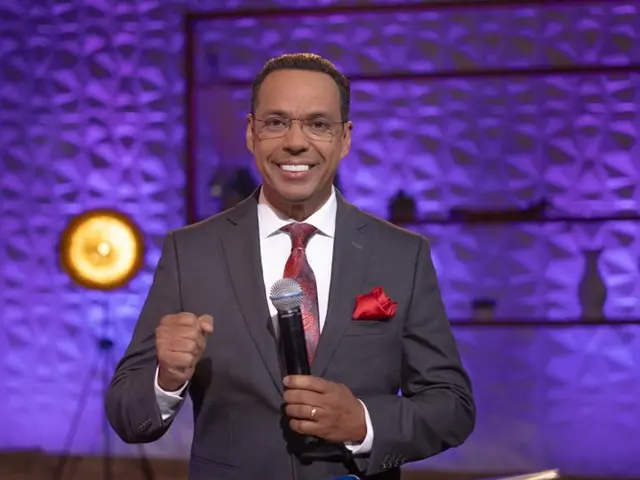 The image shows a man in a suit and tie standing on a stage, holding a microphone and wearing...