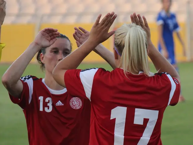 The image shows two women in red shirts and white shorts standing on a soccer field, clapping their...