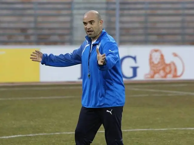 The image shows a man in a blue jacket standing on top of a soccer field, surrounded by boards with...