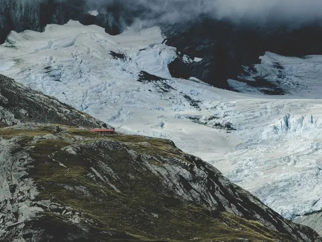 The image shows a large glacier in the middle of a mountain range, surrounded by snow-covered hills...