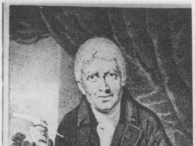 The image shows a man sitting at a table with a pen in his hand, surrounded by papers and other...