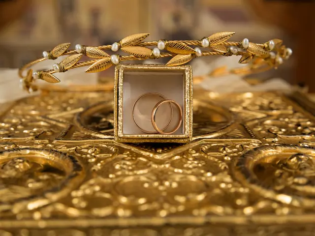 The image shows two gold wedding rings sitting atop a gold box, with a blurred background. The...