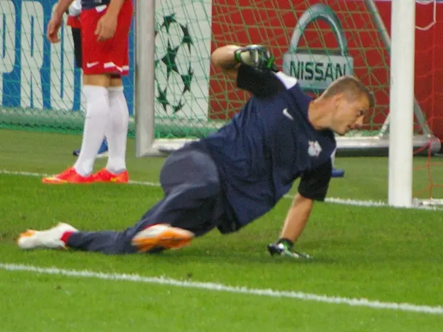 The image shows a soccer player diving to catch a ball in front of a goal, with two people standing...