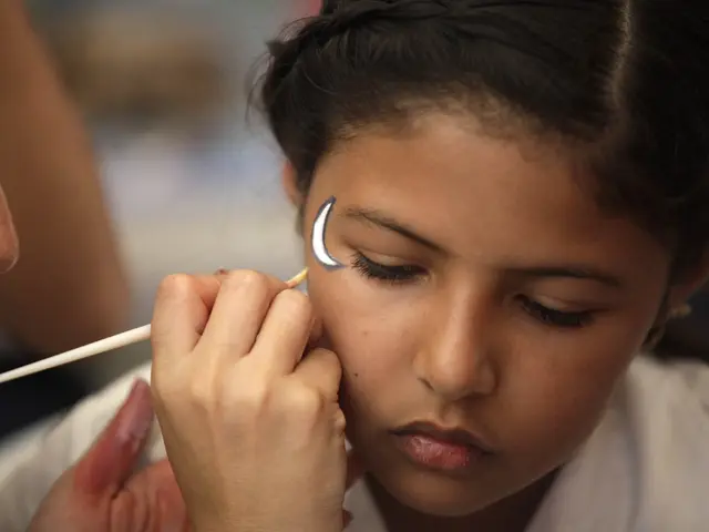 The image shows a young girl getting her make up done by a makeup artist. The makeup artist is...