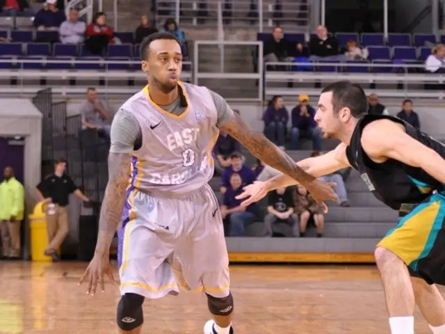 The image shows two men playing basketball on a court with a crowd of people watching. In the...