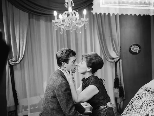 The image shows a man and woman sitting on top of a bed, embracing and kissing each other tenderly....