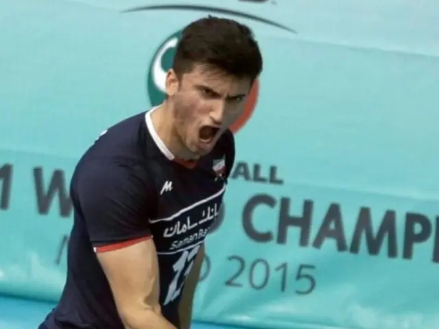 The image shows a man in a blue shirt and black shorts playing a game of handball, with a banner in...