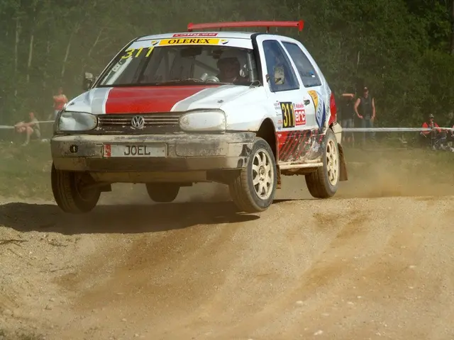 The image shows a rally car driving down a dirt road surrounded by trees. The car is adorned with...