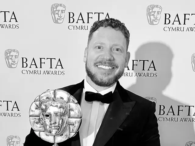 The image shows a man in a tuxedo standing and smiling while holding a BAFTA award in his hands. In...