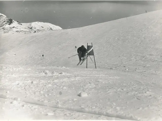The image shows an old black and white photo of a person skiing down a snowy hill, holding ski...