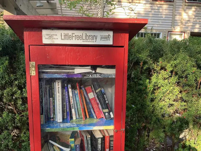 The image shows a red little free library sitting on the side of a road, surrounded by trees and a...