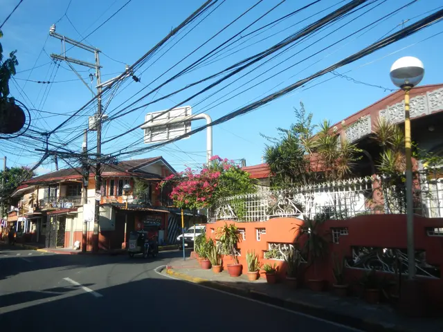 The image shows a street with power lines running down the middle of it, surrounded by buildings,...