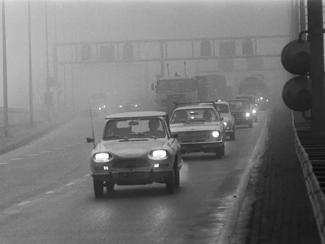 The image shows a black and white photo of cars driving down a foggy road, with a bridge in the...