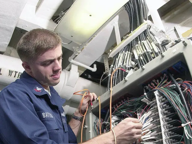 The image shows a man in a blue uniform standing and working on a computer, surrounded by wires and...
