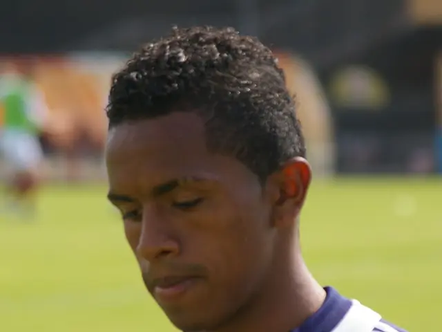 The image shows a young man in a blue t-shirt standing on top of a soccer field, with a blurred...