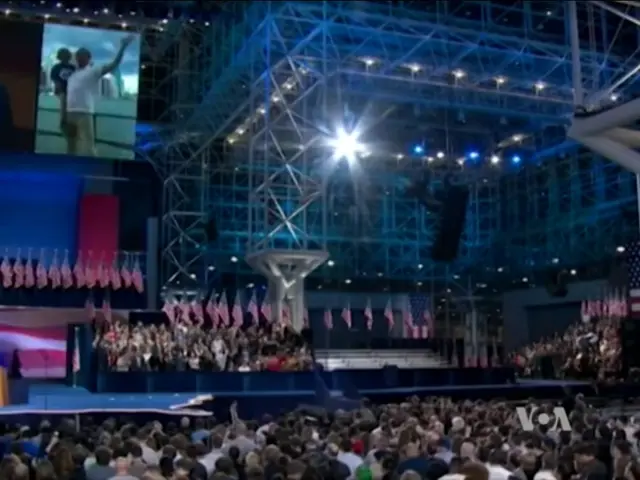 The image shows a large crowd of people sitting in front of a stage, with a podium and microphone...