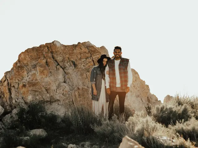 The image shows a man and woman standing on top of a rock in the desert, surrounded by lush green...