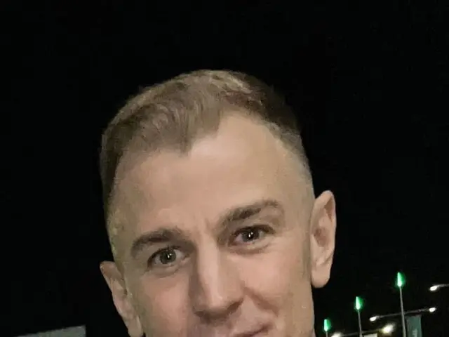 The image shows a man in a black jacket standing in front of a crowd of people, illuminated by the...