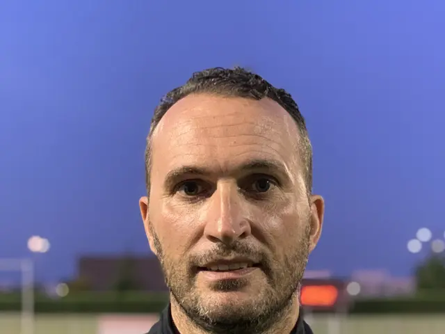 The image shows a man in a black jacket standing on top of a soccer field, with a blurred...