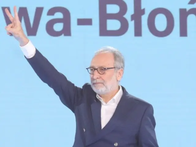 The image shows a man in a suit and tie standing on a stage, holding a microphone in his hand and...