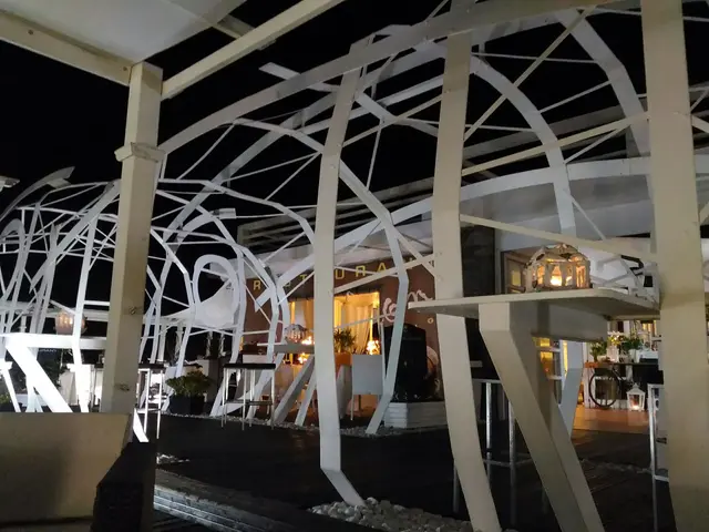 The image shows a restaurant at night with a lot of tables and chairs, illuminated by lights and...