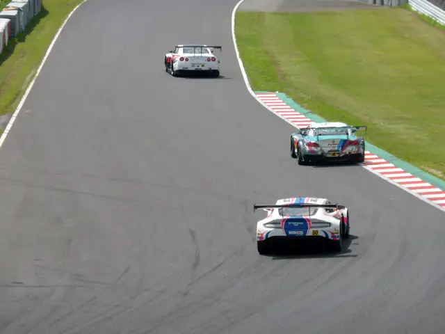 The image shows a group of cars racing down a race track, with grass on either side of the track...