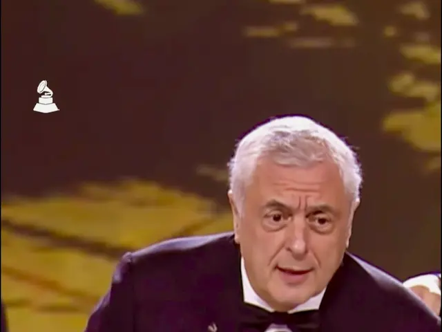 The image shows a man in a tuxedo holding a Grammy Award, with a microphone in front of him and...