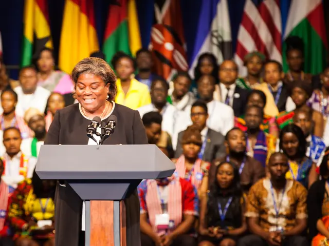 The image shows a woman standing at a podium in front of a crowd of people, with microphones on the...