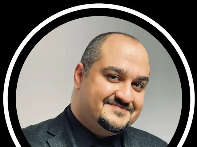 The image shows a man in a suit and tie smiling at the camera against a black background. He...