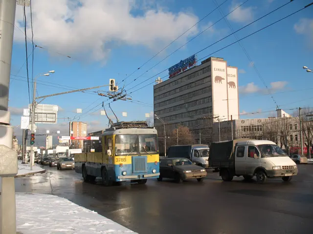 The image shows a city street with cars and trucks driving down it, surrounded by snow on the...